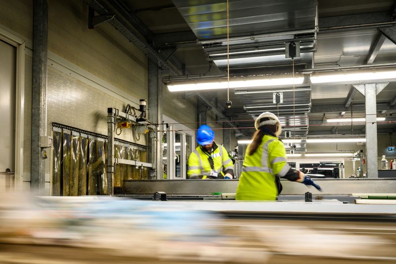 In a bustling recycling center, workers in high-visibility vests meticulously sort materials on a conveyor belt. Their focused diligence, captured with a three-quarter length portrait, underscores a commitment to environmental stewardship, ensuring quality control and responsible waste management.
