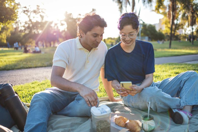 Two young friends relax in a park, using mobile phone , sharing mate tea and pleasant conversation on a sunny afternoon. The setting and their companionship create a calm and joyful ambiance.