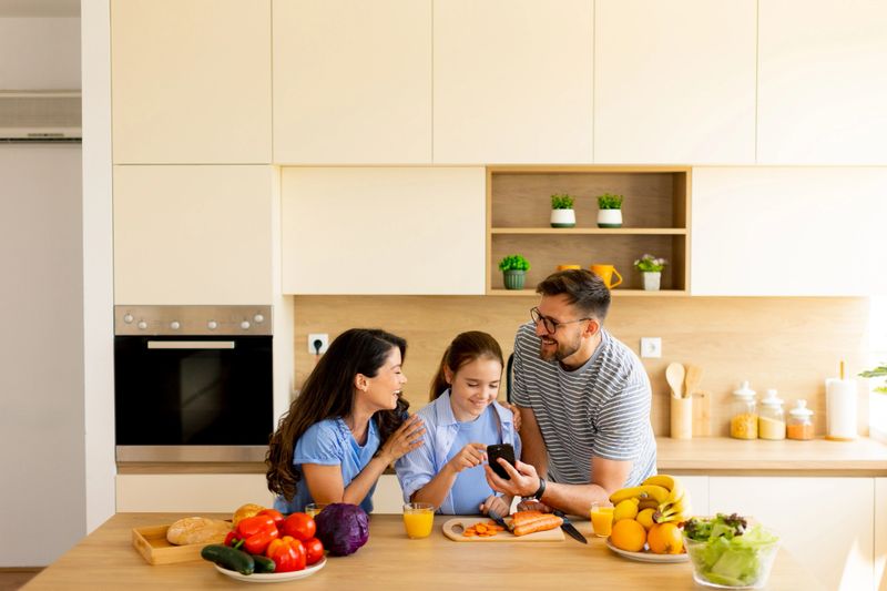 A joyful family shares a light breakfast in their spacious kitchen. They enjoy fresh fruits, veggies, and laughter while capturing a happy moment on a smartphone.