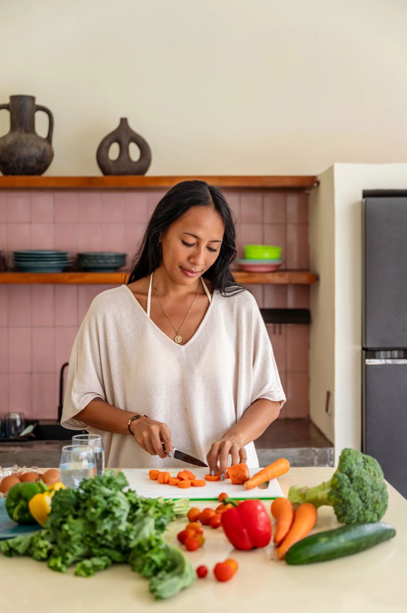 A woman is chopping fresh vegetables in her kitchen, focusing on healthy and nutritious meal preparation. The kitchen setup and vibrant ingredients create a feeling of wellness and positivity.