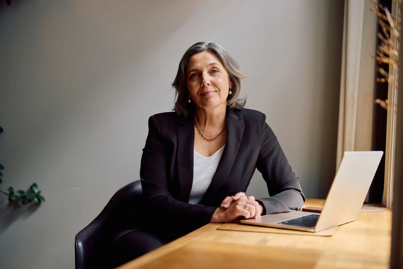 Mature professional woman in a dark blazer sitting at a wooden desk with her laptop, looking at camera confidently in a softly lit office environment with grey walls.