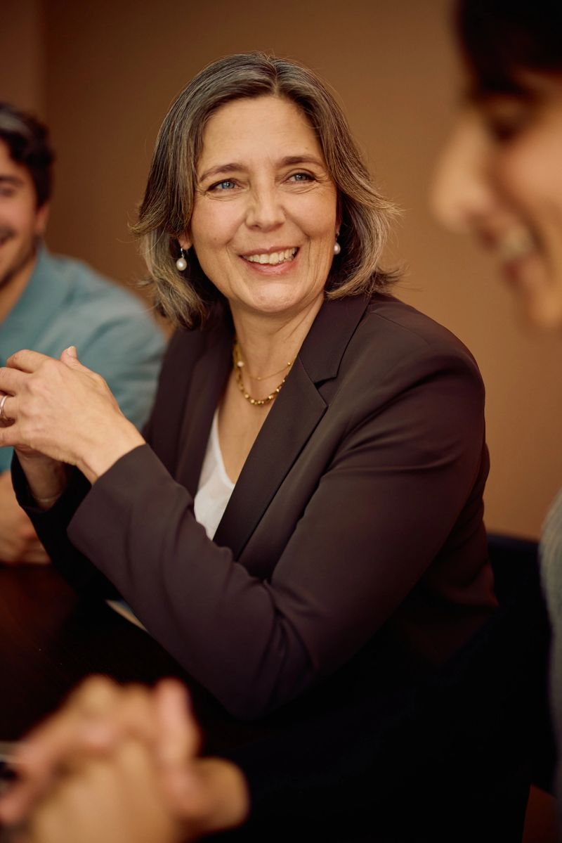 Mature professional in a brown blazer smiling warmly while seated at a meeting table, participating in a collaborative and positive workplace discussion.