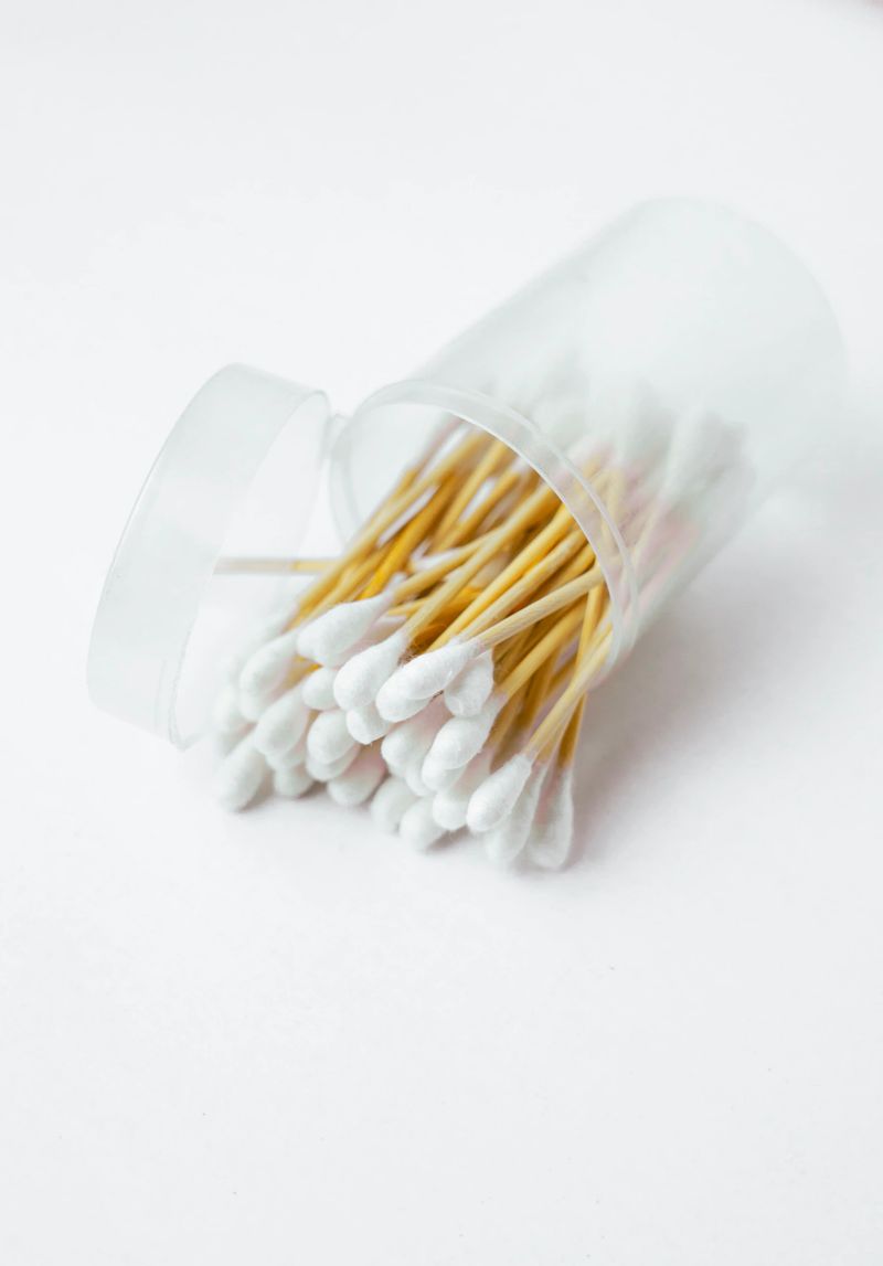 A close-up product shot of numerous cotton swabs spilled from a clear plastic container. The swabs have wooden shafts and white cotton tips. The background is plain white. This image is suitable for use in hygiene product marketing or health-related contexts.