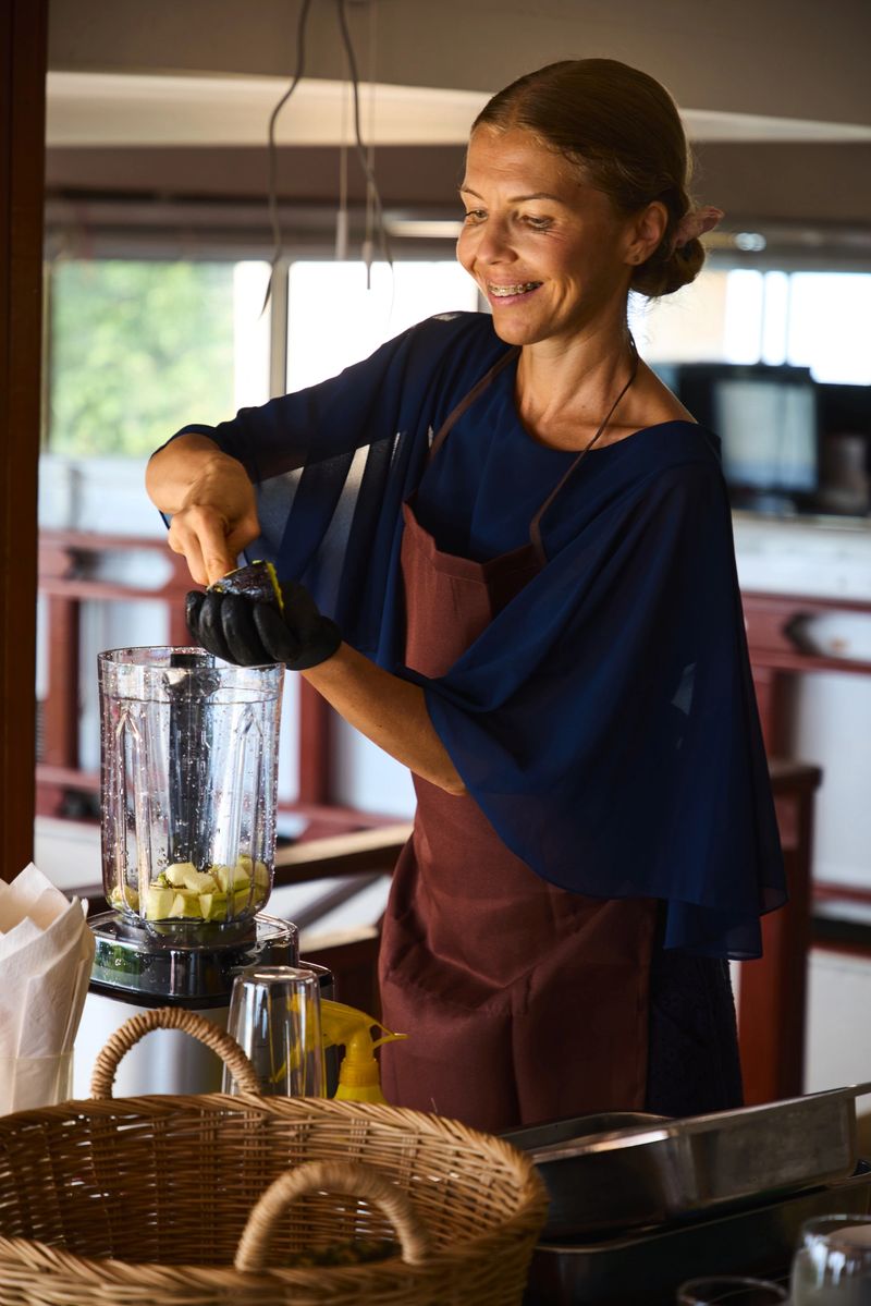Smiling woman in an apron preparing a healthy smoothie by blending fresh ingredients in a modern kitchen setting, highlighting healthy eating, culinary passion, and a joyful lifestyle.