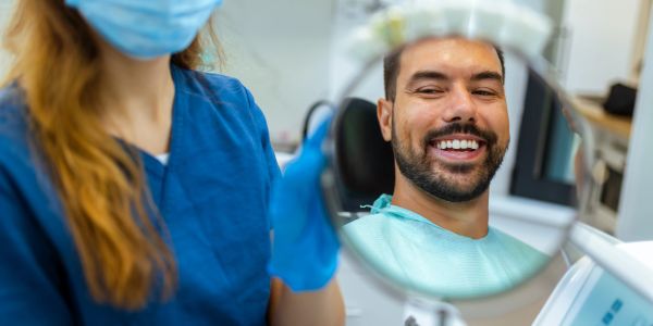 Smiling man checks his teeth in a dental mirror held by a masked dentist.