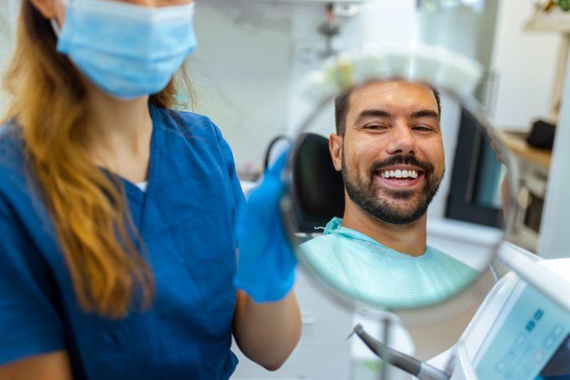 Female dentist helping male patient see his smile using a small mirror during dental checkup. Friendly and caring environment in a modern dental clinic.