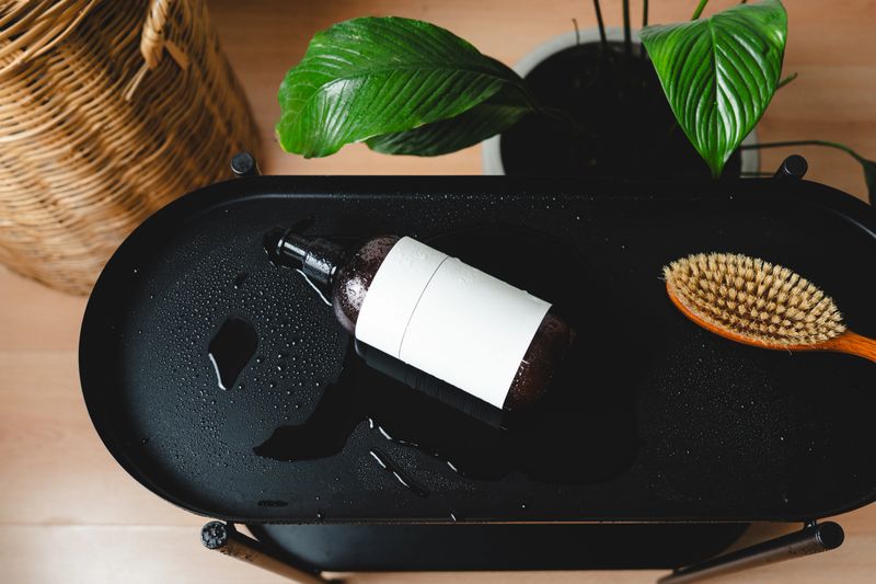 Hair care cosmetics in dark glass bottle with blank label and wooden brush on wet black surface near wicker basket and houseplant