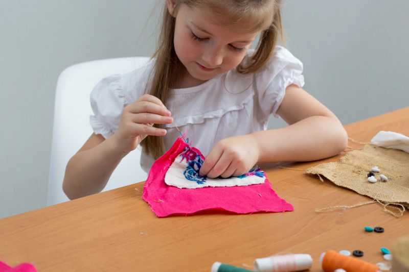Young girl working on a sewing project with colorful fabric scraps, thread spools, scissors, and buttons on a wooden table.