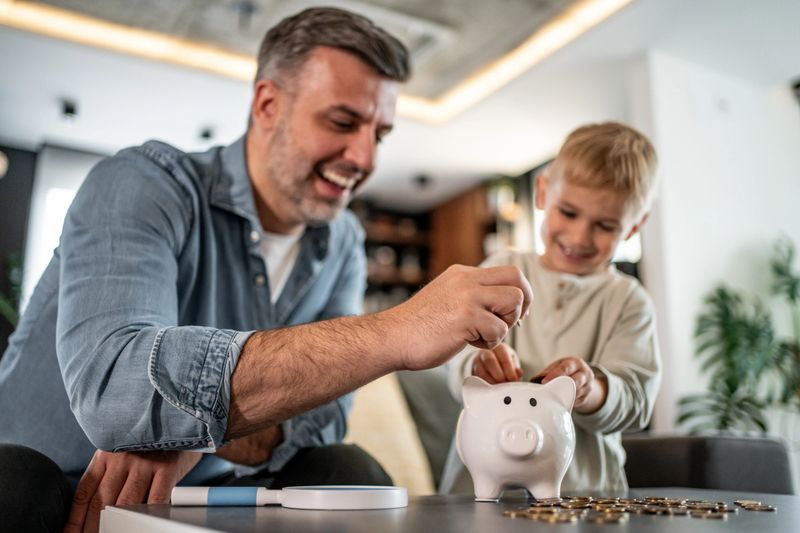 Father and son sitting together on a cozy sofa, smiling joyfully while placing coins into a cheerful piggy bank, discovering the importance of saving and personal finance