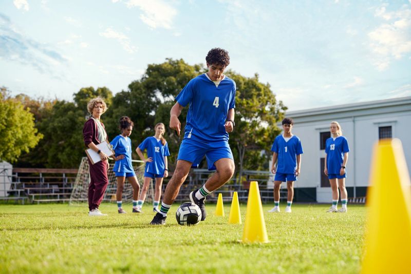Male teenager player dribbling a soccer ball through yellow cones during a training session. Female coach and teammates watching athlete practice in field. Focused athlete weaving through cones with precision during a football drill.