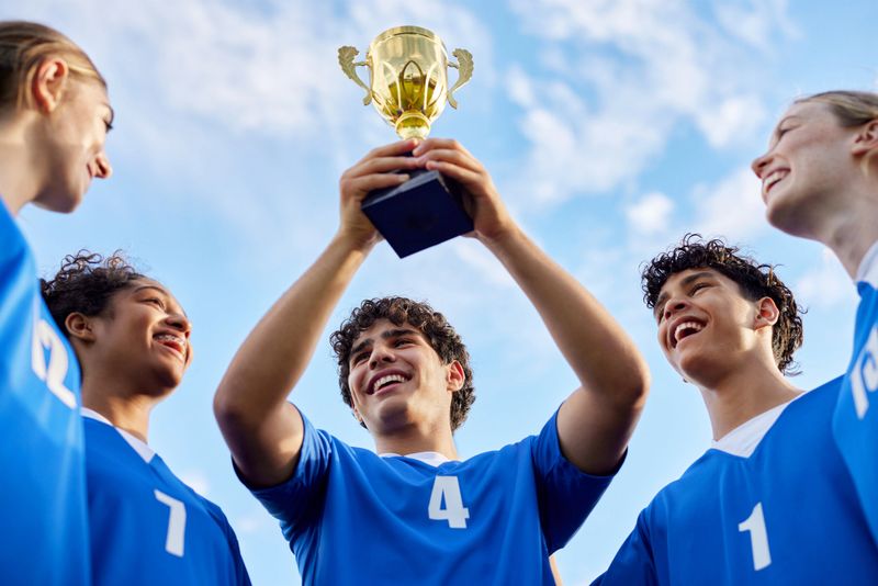 Smiling guy lifting golden trophy as teammates cheer around. Excited young multiethnic team celebrating victory on field. Joyful group of school players looking up at golden trophy in unity.