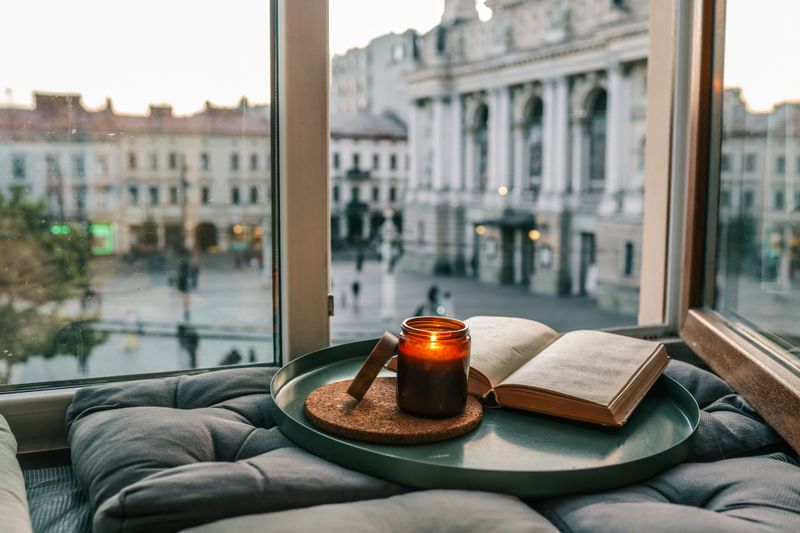 Open book and lit candle on a cozy windowsill nook with cushions, overlooking the Lviv Opera House. Warm, peaceful sunset ambiance perfect for relaxing or reading.