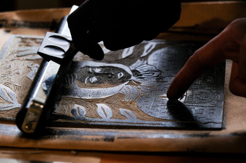 Two hands, one gloved and one bare, inspecting the details of a freshly inked linocut plate featuring a portrait design. A moment of precision before the final printing step.