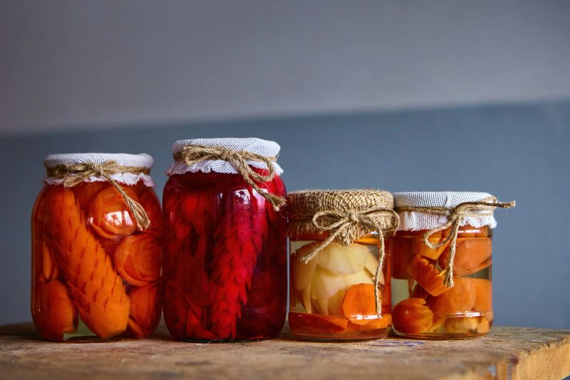 Four jars filled with pickled vegetables display vibrant colors in a rustic kitchen on a wooden table, illuminated by soft light.