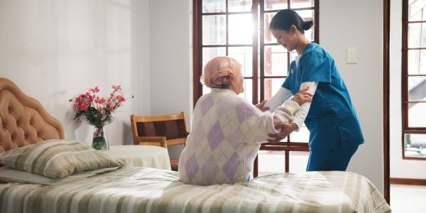 Nurse assisting elderly woman to stand beside bed.