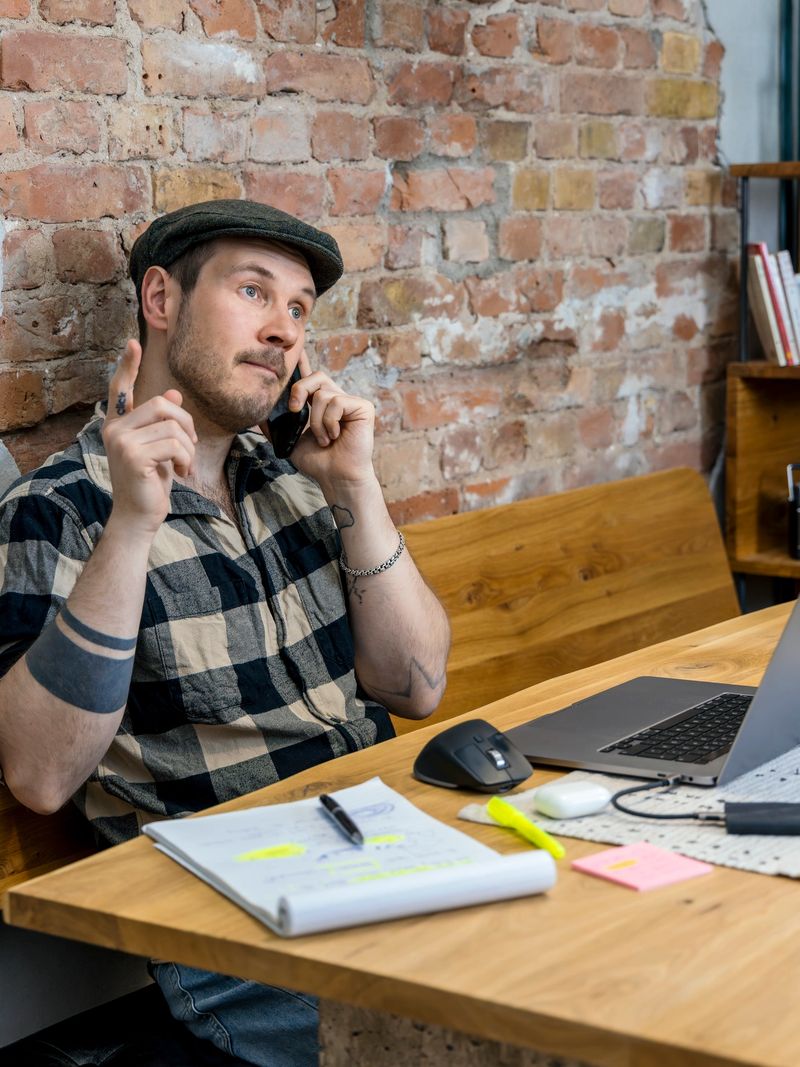 A man is seated at a wooden desk in a comfortable home office. He is talking on the phone, gesturing as he engages in a conversation.