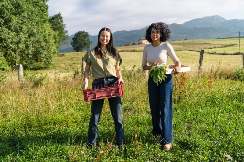 Two young women farmers are smiling at the camera while holding crates of freshly picked vegetables in a field