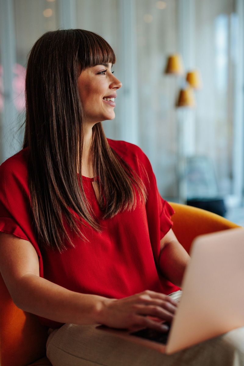 Young businesswoman sitting on an orange armchair, working on her laptop and looking away smiling