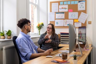 Two colleagues having a discussion in a bright office with charts on a bulletin board.