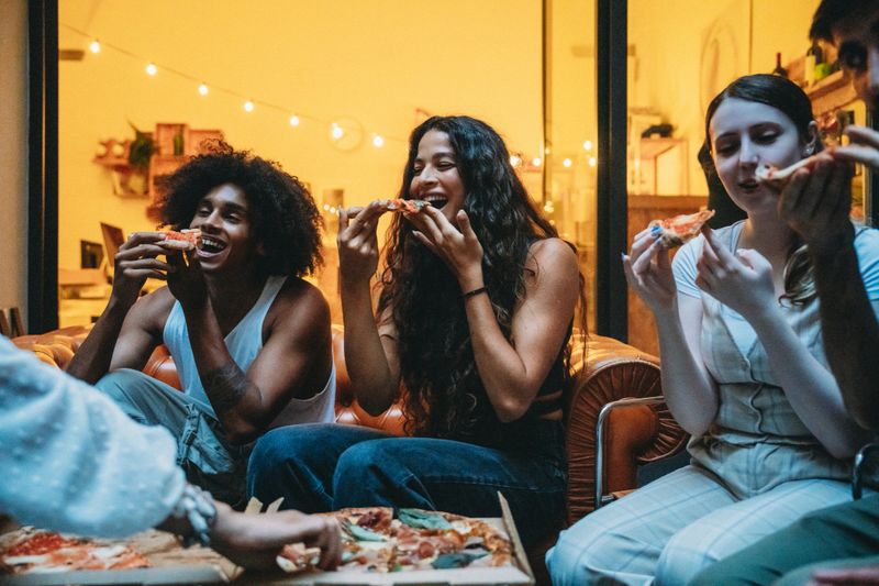 Group of cheerful young friends sharing delicious pizza, laughing heartily and enjoying a lively evening filled with fun and connection