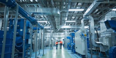 Two workers walk inside a large industrial facility with extensive piping and machinery.