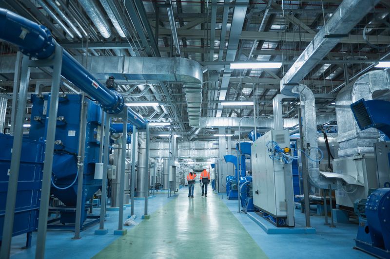 Two engineers wearing safety helmets and high-visibility orange jackets walk through a high-tech industrial facility, with discuss job. The environment is filled with advanced machinery, large metal pipes, and complex ventilation systems.
