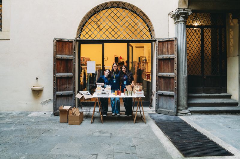 Three young adult female volunteers smiling behind a table selling goods at a charity event in Florence, Italy