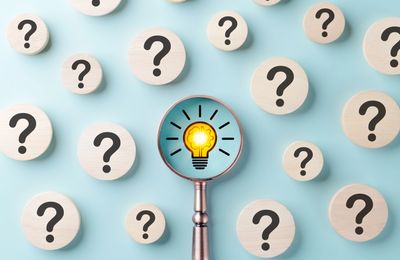 Magnifying glass reveals a glowing light bulb among question marks.