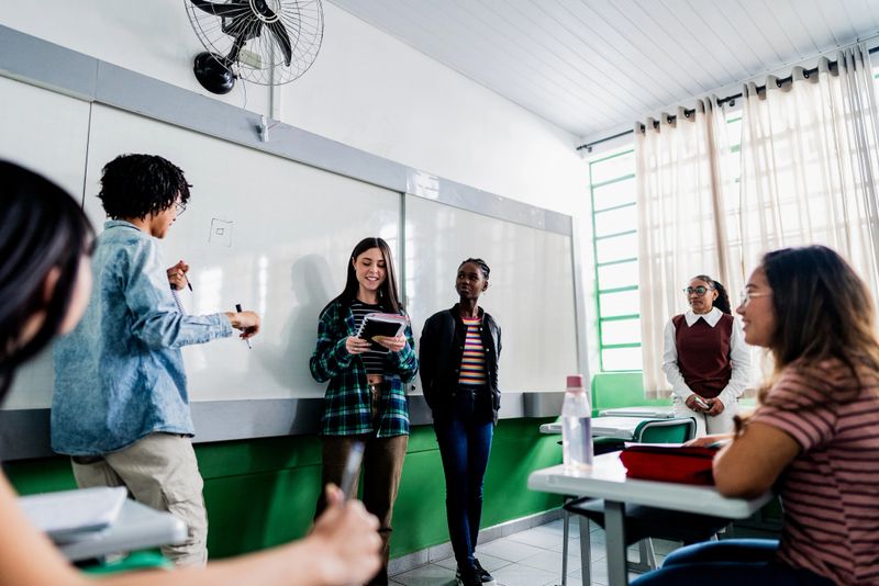 Students stand in front of the whiteboard, holding materials and presenting a school project to the class. Their classmates watch attentively from their desks. The scene highlights engagement, collaboration, and the development of communication skills in an educational environment