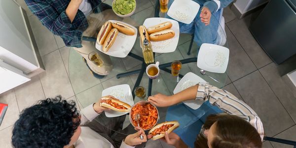 Four people enjoying hot dogs with fresh toppings around a glass table.