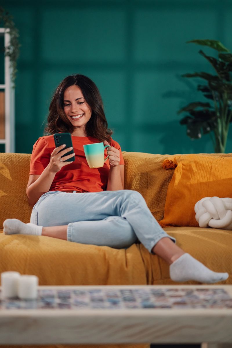Smiling young woman relaxing on a comfortable sofa at home, enjoying a cup of coffee and browsing her smartphone
