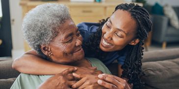 A young woman embracing an elderly woman with warm smiles on a couch.