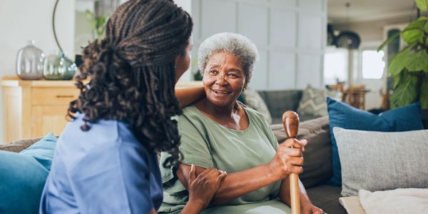 Caregiver smiling at elderly woman holding a walking cane at home.