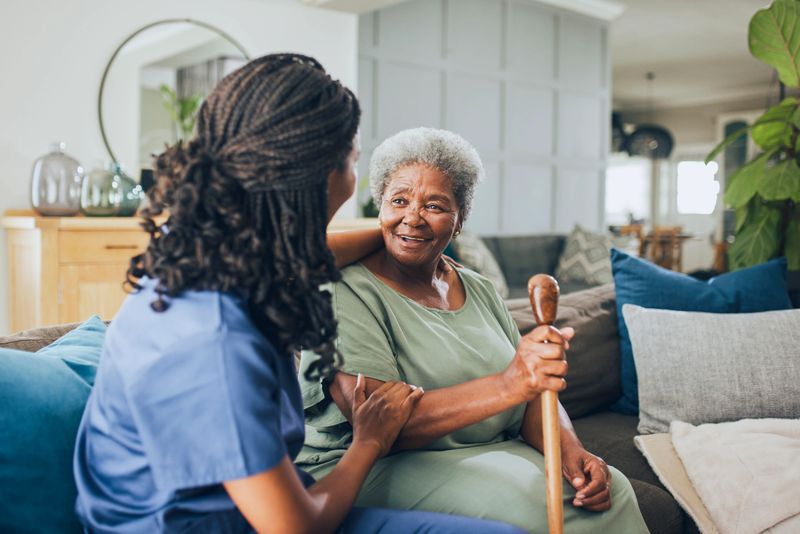 Portrait of a young female nurse having a happy conversation and comfortingly holding the arm of a senior woman who is holding a walking cane and sitting on a couch at home.