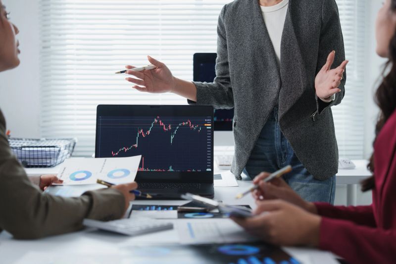 Businesswomen analyzing financial charts on a laptop during an office meeting, discussing investment strategies and market trends
