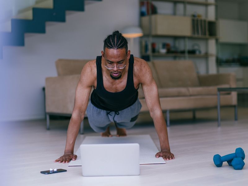 Strong African American man performing push ups in front of laptop following online fitness program on living room floor.