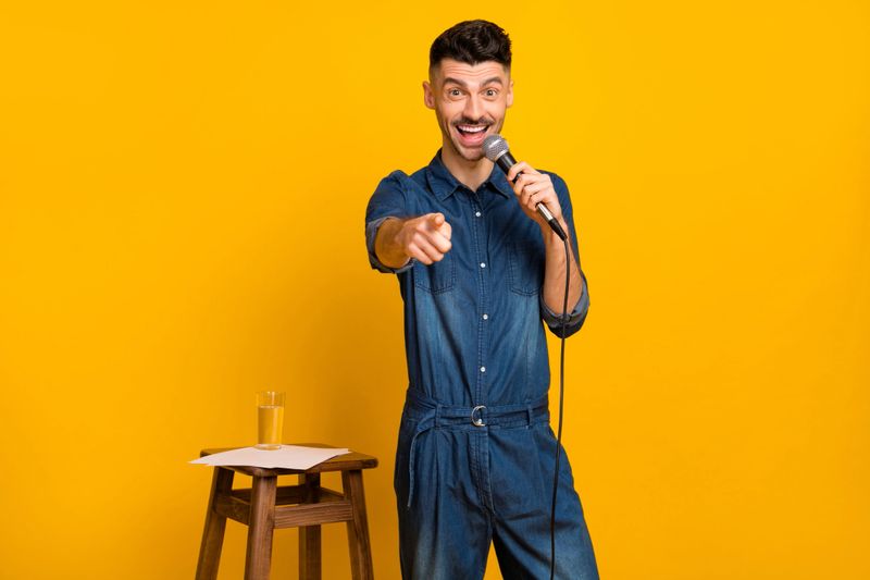 Photo of handsome brunette young man comedian hold mic point finger you isolated on yellow color background