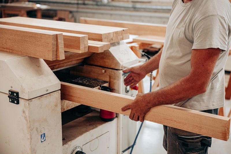 A carpenter feeds an oak beam into a woodworking planer machine inside a carpentry workshop, carefully smoothing the surface for precision furniture construction
