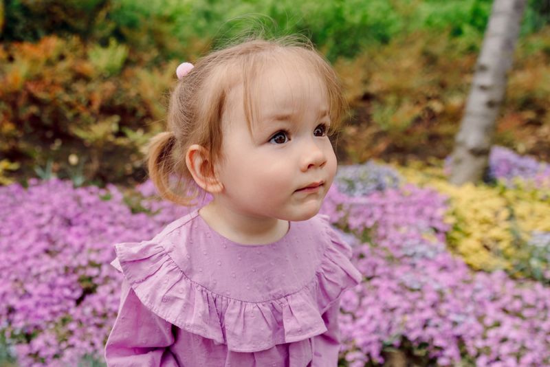 Adorable little girl in purple dress looking up with curiosity in a blooming spring garden.