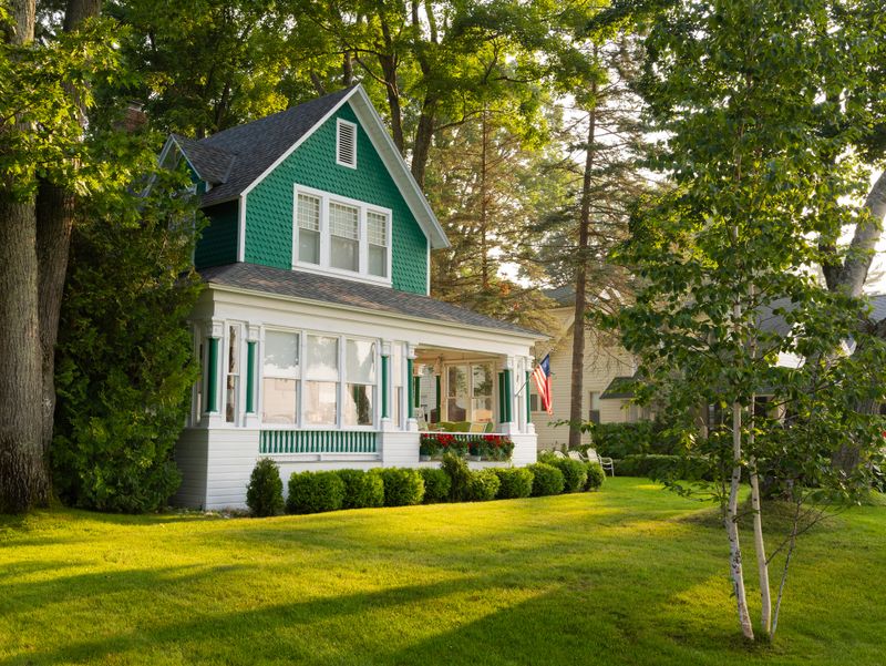 With an American flag proudly displayed, a corner view of a Lake Michigan seasonal summer cottage that is painted green and white. Built in the late 1800's, the house is surrounded by a lush lawn, trees and bushes. It is the perfect escape for vacation, rest and relaxation.