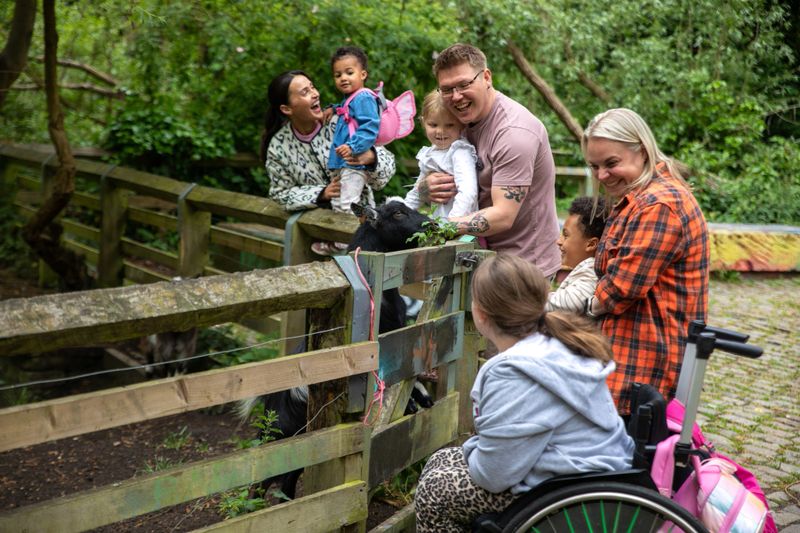 A medium close up side view of a familes and their children visiting some goats grazing on a field on a family day out to a community charity farm in Newcastle upon Tyne in the North East of England. One of the children is disabled and a wheelchair user and is having a fun day with her parents and her little sister.Videos available for this scenario