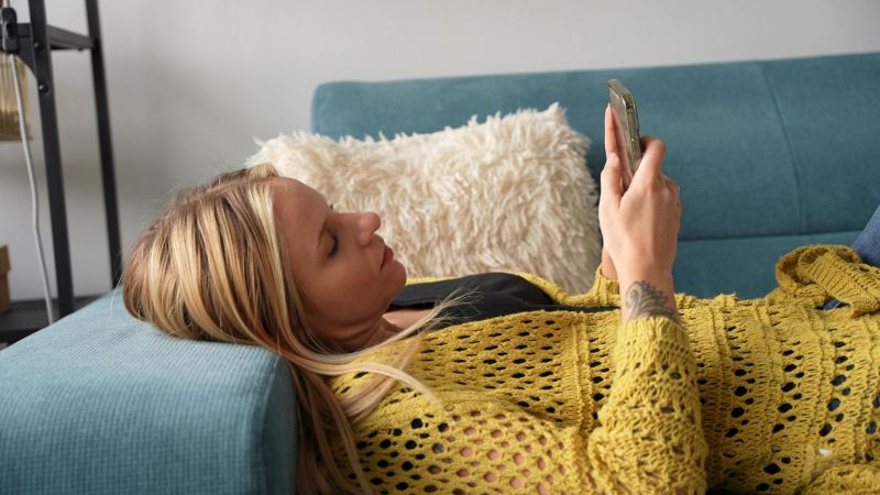 Relaxed Young Woman Lounging on the Sofa, Scrolling Through her Smartphone, Enjoying Leisure Time at Home While Connecting with Friends and Browsing the Latest Social Media Updates