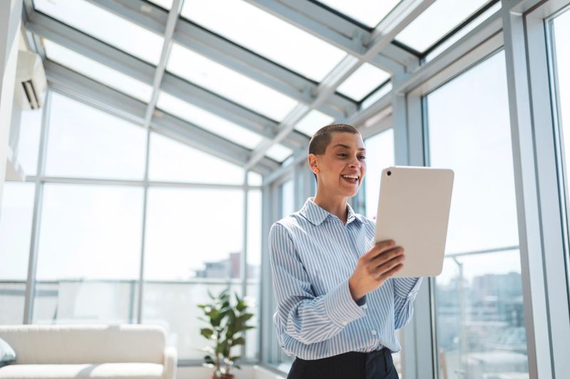 Professional individual working with a digital tablet in a bright, glass-panelled office space. The environment suggests a modern, airy workspace, emphasizing productivity and engagement with digital technology.