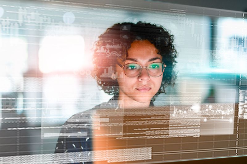 A professional focuses intently on data visible on a futuristic transparent screen, enhanced with overlay graphics under ambient light, symbolizing modern technology, analytics, and digital innovation in a professional setting.