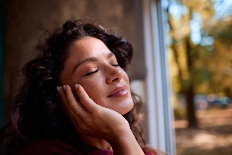 Mid adult woman with curly hair and closed eyes gently holds her face, finding tranquility and enjoying a peaceful moment by the window with a blurred autumnal view in the background
