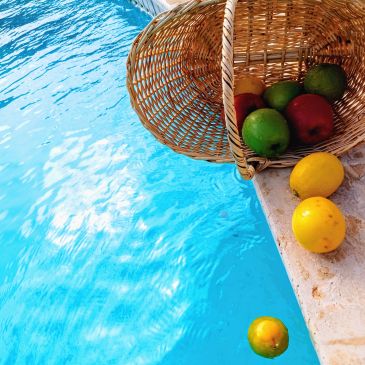 A basket with fruits tipped over beside a swimming pool with some fruits fallen inside and outside.