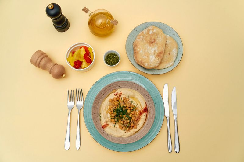 A Vibrant Cuisine Spread Featuring Hummus, Pita Bread, Olive Oil, and Fresh Ingredients Arranged Beautifully Against a Soft Yellow Background