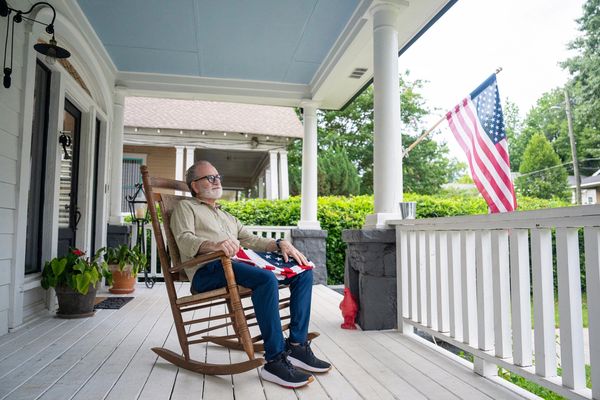An elderly man sitting on a porch rocking chair holding an American flag.