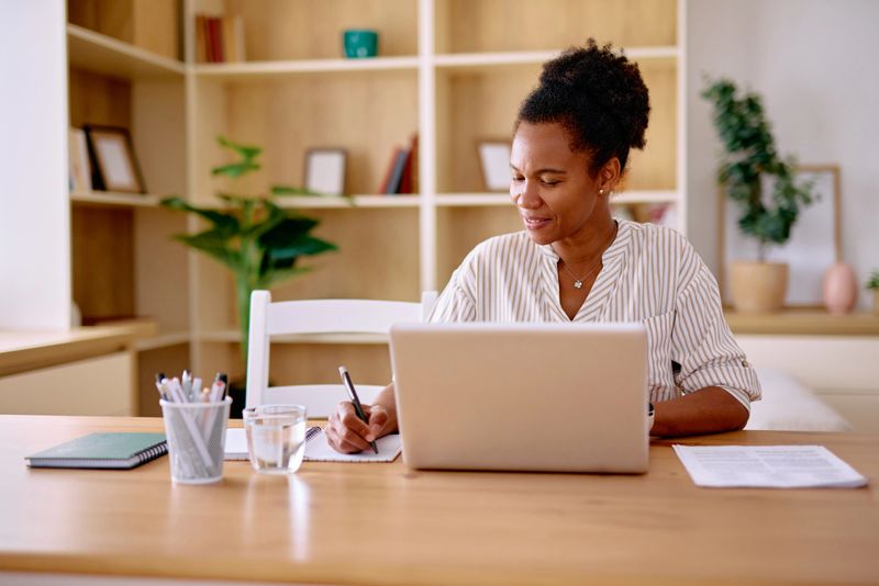 Mid adult African American woman taking notes while working on a laptop in a cozy home office. Surrounded by plants and natural light, focusing on productivity and organization