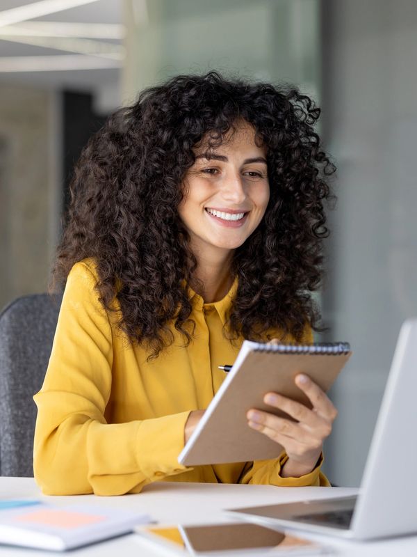 A smiling woman with long, dark, curly hair, wearing a yellow shirt, holding a notebook and a pen